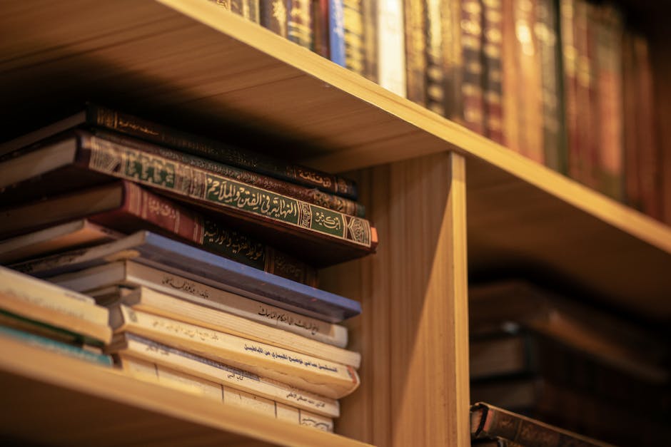 Foto naskah kitab kuning klasik yang terbuka di atas meja kayu dengan pencahayaan hangat, dikelilingi oleh tumpukan literatur hadits dan tafsir di sebuah perpustakaan Islam tradisional yang tenang.
