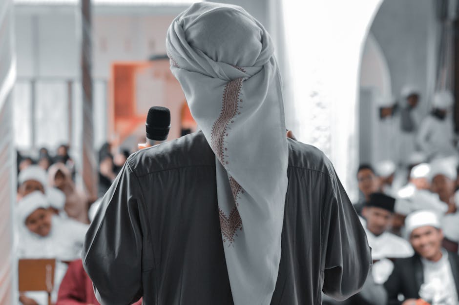 Foto sebuah mushaf Al-Quran terbuka di atas meja kayu di dalam perpustakaan klasik dengan cahaya lampu temaram yang tenang, dikelilingi oleh tumpukan kitab-kitab turats kuno.