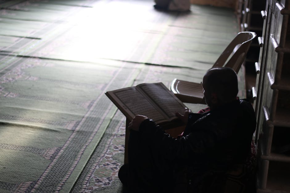 Foto sebuah naskah kuno kitab kuning klasik yang terbuka di atas meja kayu dengan pencahayaan hangat, dikelilingi oleh rak buku perpustakaan Islam yang penuh dengan kitab-kitab teologi dan tafsir.