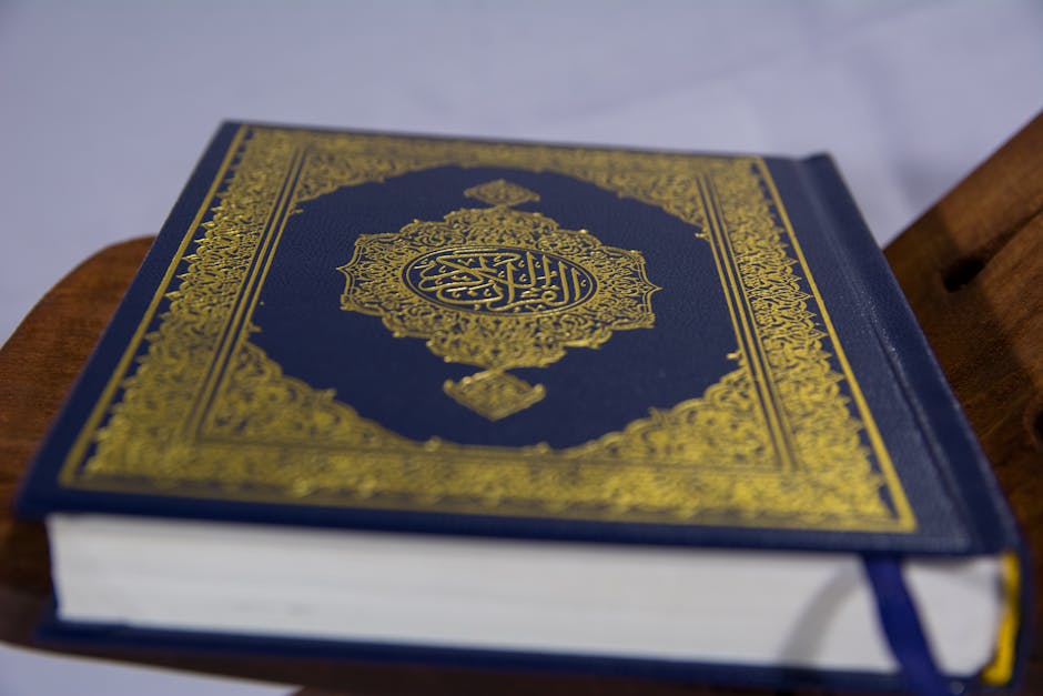 Foto sebuah naskah klasik kitab kuning beraliran Asy'ariyah yang diletakkan di atas meja kayu tua dengan pencahayaan hangat dari lampu gantung di dalam sebuah perpustakaan Islam tradisional yang penuh dengan jajaran kitab-kitab tebal di latar belakang.