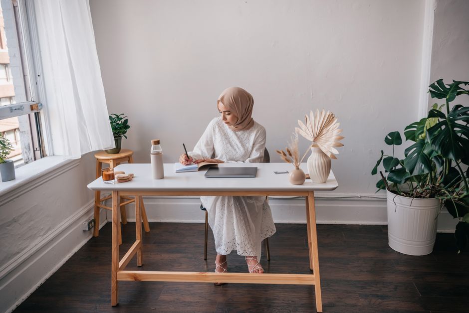 Ilustrasi seorang pemikir Muslim paruh baya yang sedang menulis catatan di meja kayu dengan latar belakang rak buku klasik yang penuh, menciptakan suasana reflektif, tenang, dan penuh kedalaman ilmu.
