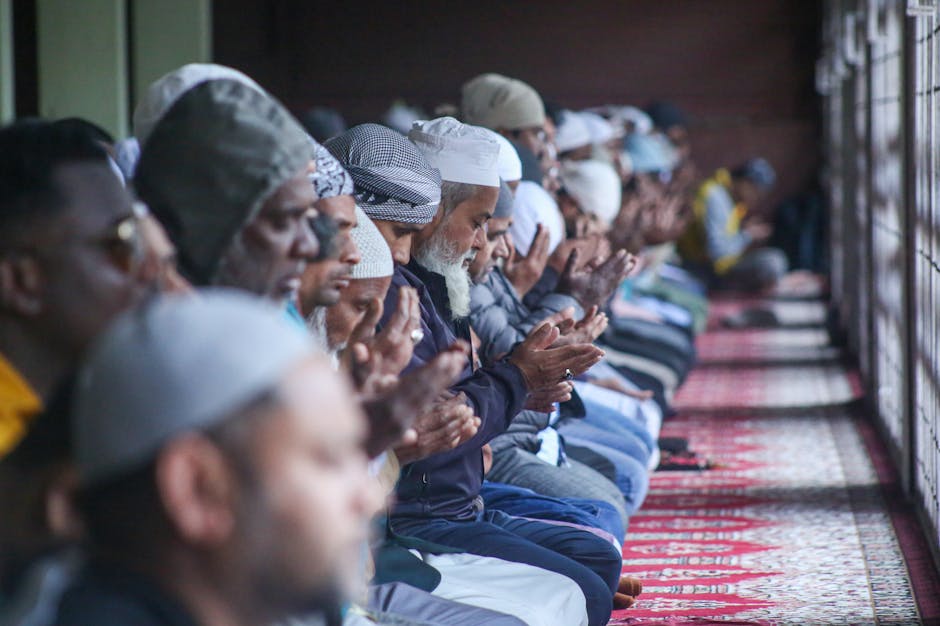 Foto sebuah naskah klasik kitab kuning yang terbuka di atas meja kayu tua dengan pencahayaan hangat, di sampingnya terletak tasbih dan sajadah tipis di dalam sebuah masjid dengan arsitektur klasik yang tenang.