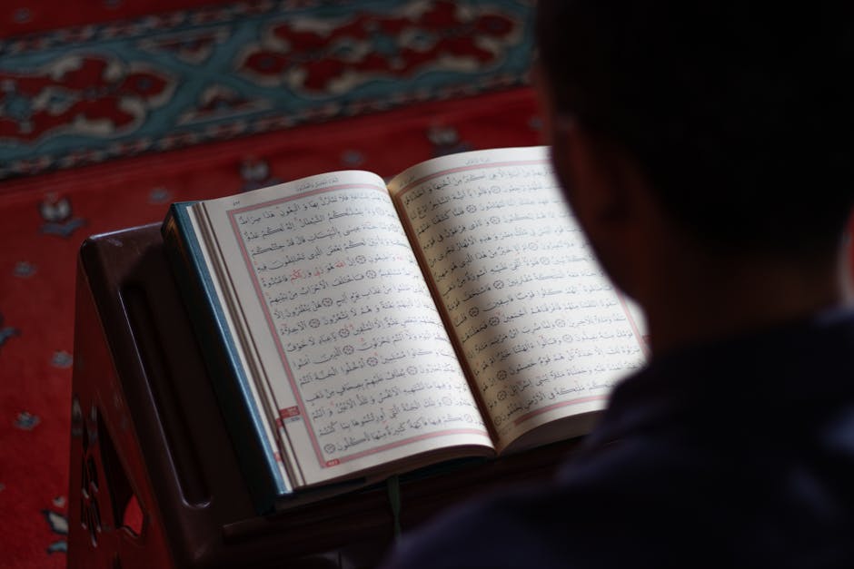 Foto sebuah mushaf Al-Quran terbuka dengan pencahayaan lembut di sebuah perpustakaan klasik yang penuh dengan deretan kitab-kitab turats, menciptakan suasana keilmuan yang tenang dan berwibawa.