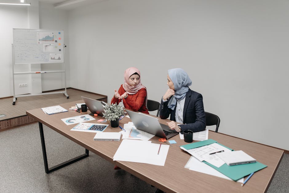 Ilustrasi seorang Muslimah yang sedang menulis di meja kerja dengan latar belakang perpustakaan dan jendela yang menampilkan pemandangan kota modern, melambangkan perpaduan intelektualitas, tradisi, dan kemajuan zaman.