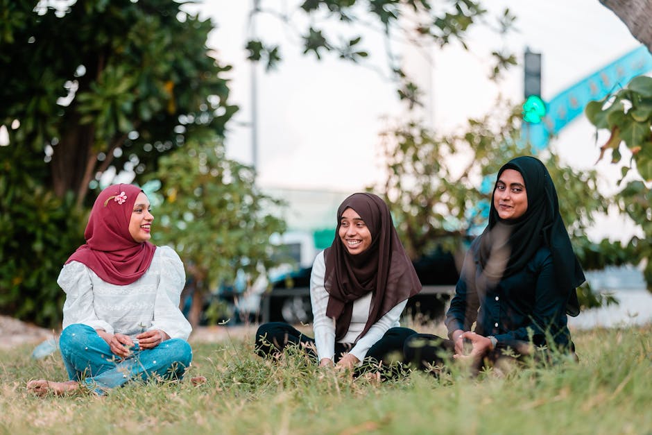 Seorang pemuda Muslim mengenakan hoodie modern sedang duduk santai di taman kota yang hijau, memegang buku catatan dan tersenyum tenang sambil melihat ke arah langit, dengan suasana cahaya sore yang hangat (golden hour).