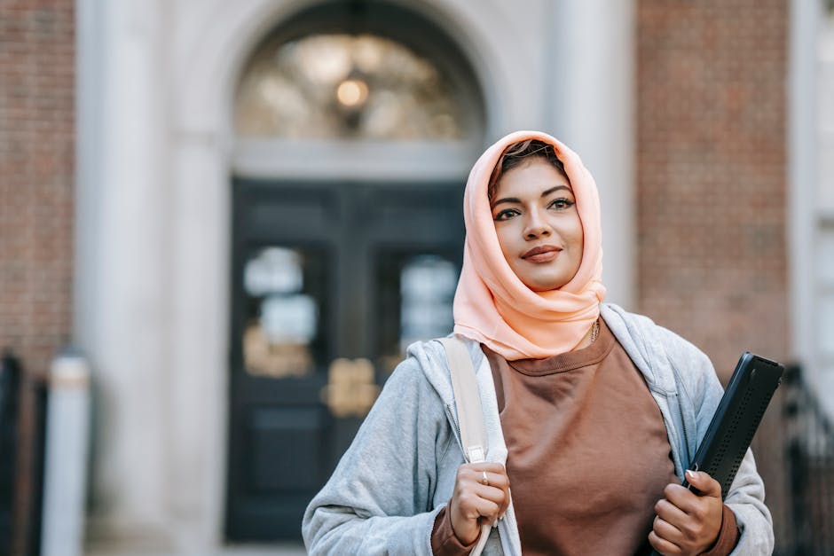 Foto estetik seorang anak muda Muslim sedang duduk santai di coffee shop modern, memakai hoodie dan headphone, sambil memegang buku catatan kecil dengan latar belakang jendela kota yang senja.