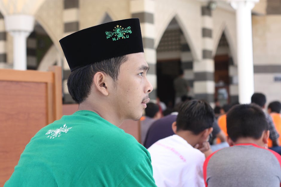 Seorang pemuda Muslim mengenakan pakaian kasual modern sedang duduk santai di sebuah taman kota yang estetik, memegang buku catatan dan tersenyum tenang sambil melihat pemandangan, mencerminkan kedamaian pikiran di tengah kesibukan dunia.