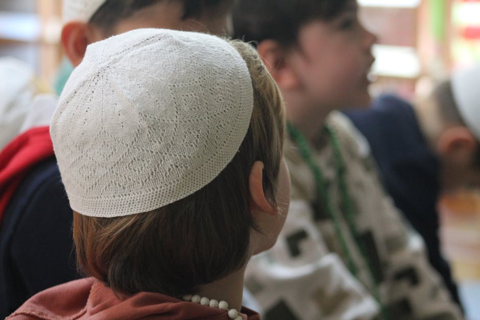 Seorang anak muda Muslim mengenakan hoodie dan sneakers sedang duduk santai di sebuah taman kota yang modern sambil memegang buku catatan, wajahnya terlihat tenang dengan latar belakang gedung-gedung perkotaan yang estetik.