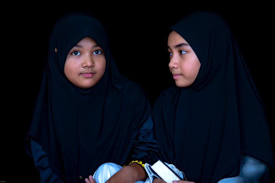 Seorang pemuda Muslim mengenakan hoodie modern sedang duduk santai di taman kota yang asri sambil memegang buku, terlihat tenang dan damai dengan pemandangan gedung-gedung tinggi yang estetik di latar belakang.