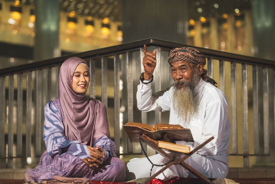 Seorang pemuda Muslim mengenakan jaket kasual modern sedang duduk santai di sudut kafe estetik, memegang secangkir kopi hangat sambil tersenyum menatap layar laptopnya dengan suasana yang tenang dan hangat.