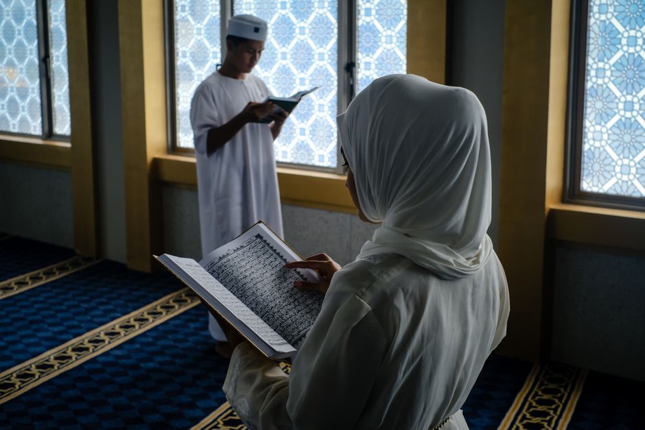 Seorang pemuda Muslim mengenakan pakaian kasual modern sedang duduk santai di sudut kafe yang estetik, memegang segelas kopi hangat sambil tersenyum menatap layar laptopnya dengan ekspresi tenang dan damai.