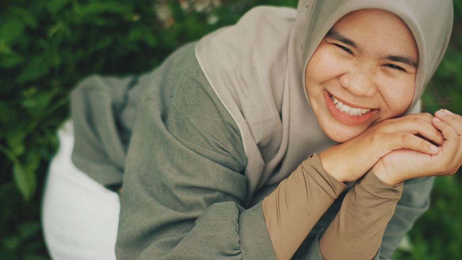 Seorang pemuda Muslim modern mengenakan hoodie kasual sedang duduk santai di sudut kafe estetik, memegang secangkir kopi hangat sambil tersenyum menatap layar smartphone-nya dengan tenang, mencerminkan kedamaian pikiran di tengah kesibukan urban.