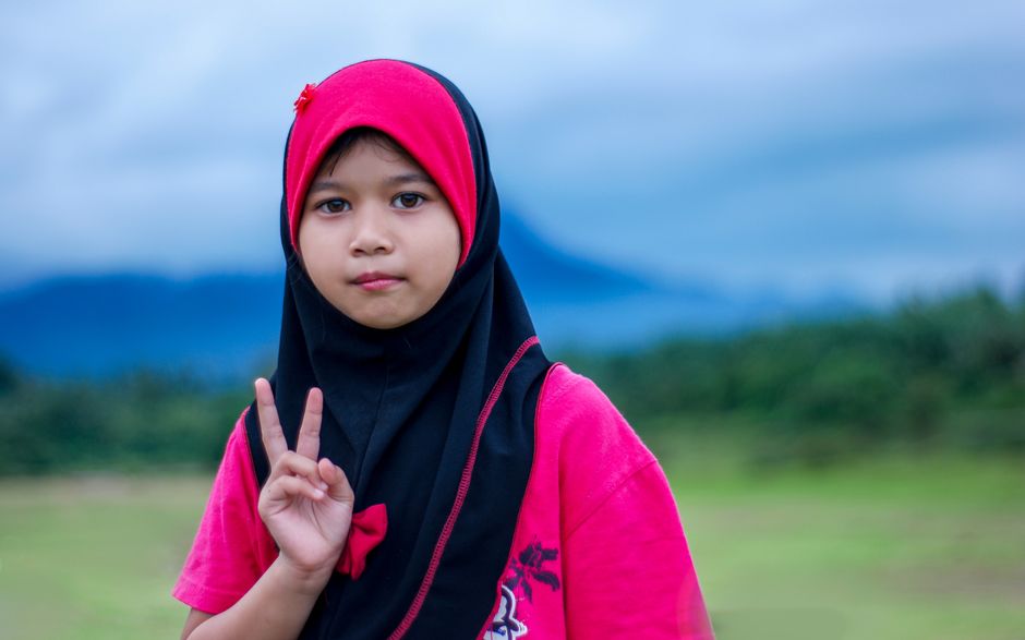 Anak muda Muslim modern sedang duduk santai di sebuah kafe estetik, memegang cangkir kopi sambil tersenyum melihat laptop, dengan suasana ruangan yang hangat dan dipenuhi tanaman hijau.