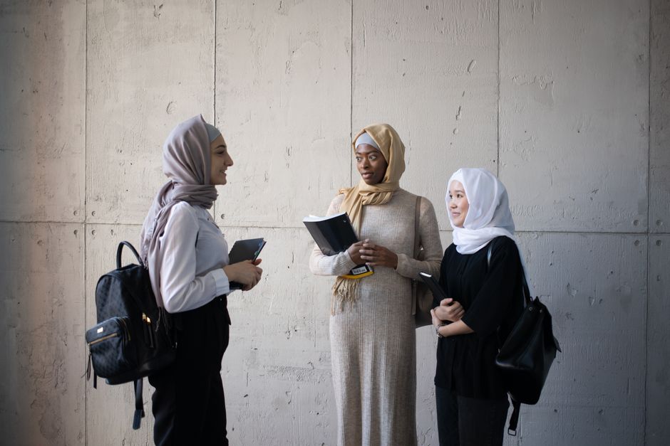 Dua orang pemuda Muslim modern sedang duduk santai di area outdoor sebuah kafe minimalis, salah satunya memegang tablet dan yang lainnya tersenyum sambil mendengarkan, suasana hangat dengan pencahayaan sore hari yang estetik.
