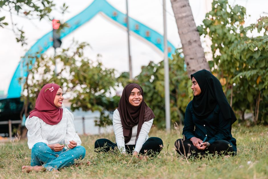 Seorang pemuda Muslim kasual mengenakan hoodie modern sedang duduk santai di sudut kafe yang estetik dan tenang, memegang secangkir kopi hangat sambil tersenyum menatap layar ponselnya dengan ekspresi damai tanpa beban.
