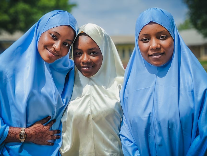Tiga orang anak muda Muslim, dua perempuan berhijab kasual modern dan satu laki-laki, sedang duduk santai sambil berdiskusi hangat di sebuah kafe estetik dengan dekorasi tanaman hijau dan pencahayaan hangat yang ramah lingkungan.