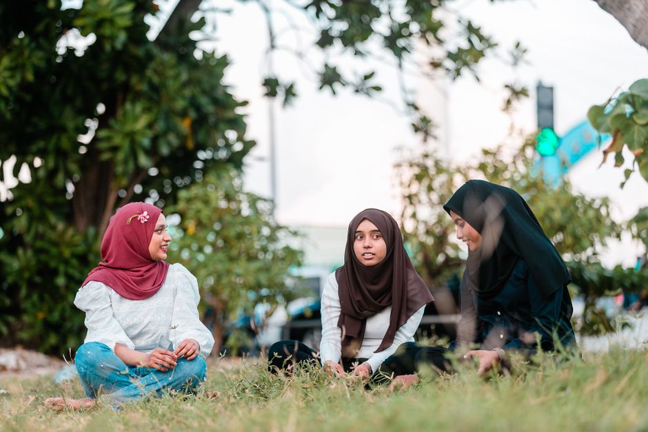 Seorang pemuda Muslim Gen Z sedang duduk santai di sudut kafe modern yang hangat dan estetik, memegang cangkir kopi sambil memandang ke luar jendela dengan ekspresi wajah yang tenang dan damai, dengan laptop serta buku catatan terbuka di atas meja.