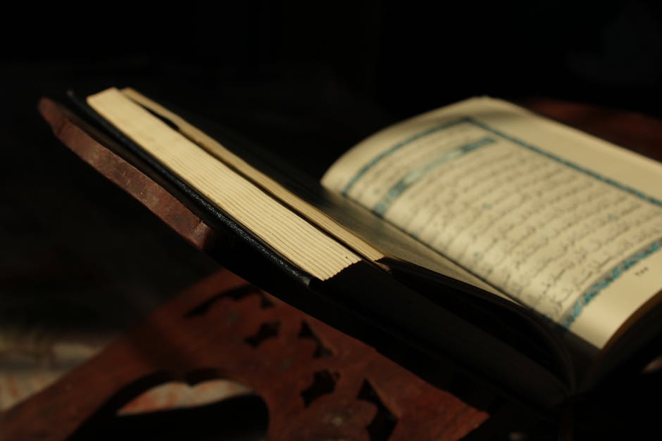 Foto sebuah mushaf Al-Quran terbuka yang diletakkan di atas meja kayu di dalam perpustakaan masjid yang tenang, dengan cahaya matahari pagi yang masuk melalui jendela memberikan kesan sakral dan ilmiah.