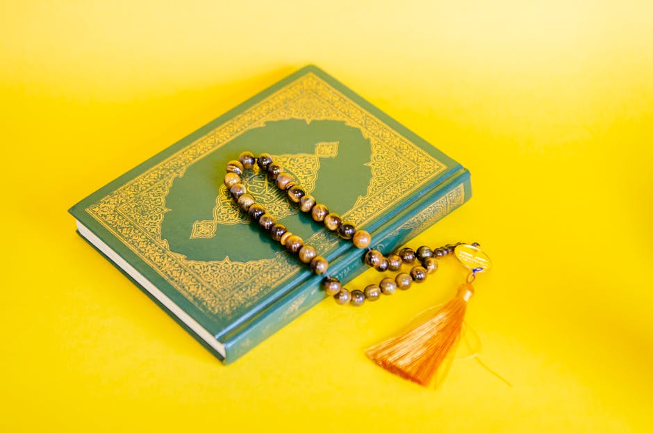 Foto sebuah mushaf Al-Quran tua yang terbuka di atas meja kayu di dalam perpustakaan klasik dengan pencahayaan hangat yang masuk dari jendela masjid, menciptakan suasana spiritual yang mendalam dan tenang.