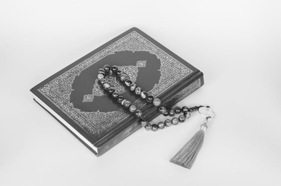 Foto sebuah mushaf Al-Quran kuno yang terbuka di atas meja kayu berukir di dalam perpustakaan klasik dengan pencahayaan hangat, dikelilingi oleh tumpukan kitab-kitab kuning karya ulama salaf.