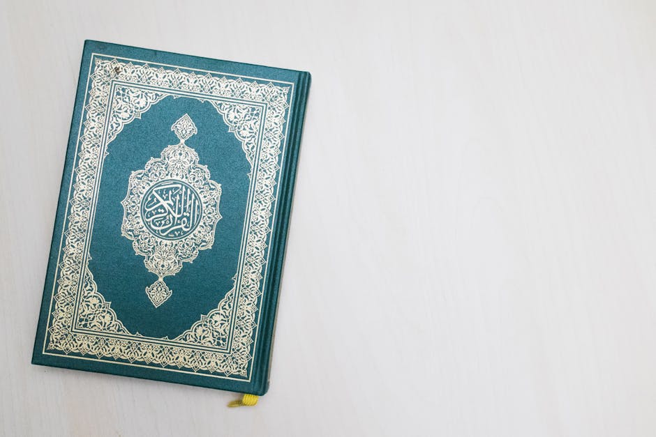 Foto sebuah naskah kuno kitab hadits dengan tulisan kaligrafi Arab yang indah, diletakkan di atas meja kayu tua di dalam perpustakaan klasik yang diterangi cahaya lampu temaram, memberikan kesan kedalaman ilmu dan kewibawaan tradisi intelektual Islam.