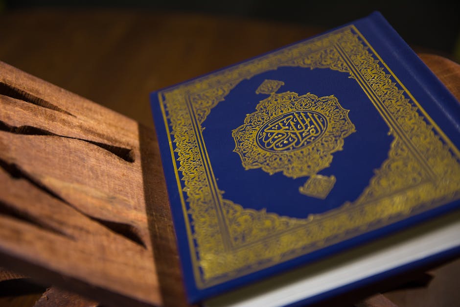 Foto sebuah mushaf Al-Quran terbuka dengan pencahayaan lembut di dalam sebuah perpustakaan klasik Islam, dikelilingi oleh tumpukan kitab-kitab turats yang tebal, menciptakan suasana keilmuan yang mendalam dan tenang.