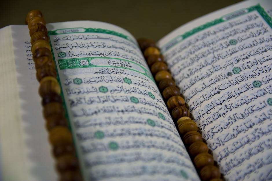 Foto sebuah naskah hadis kuno dengan tulisan kaligrafi Arab yang indah di atas meja kayu tua, dikelilingi oleh tumpukan kitab-kitab turats di dalam sebuah perpustakaan Islam klasik yang diterangi cahaya temaram.